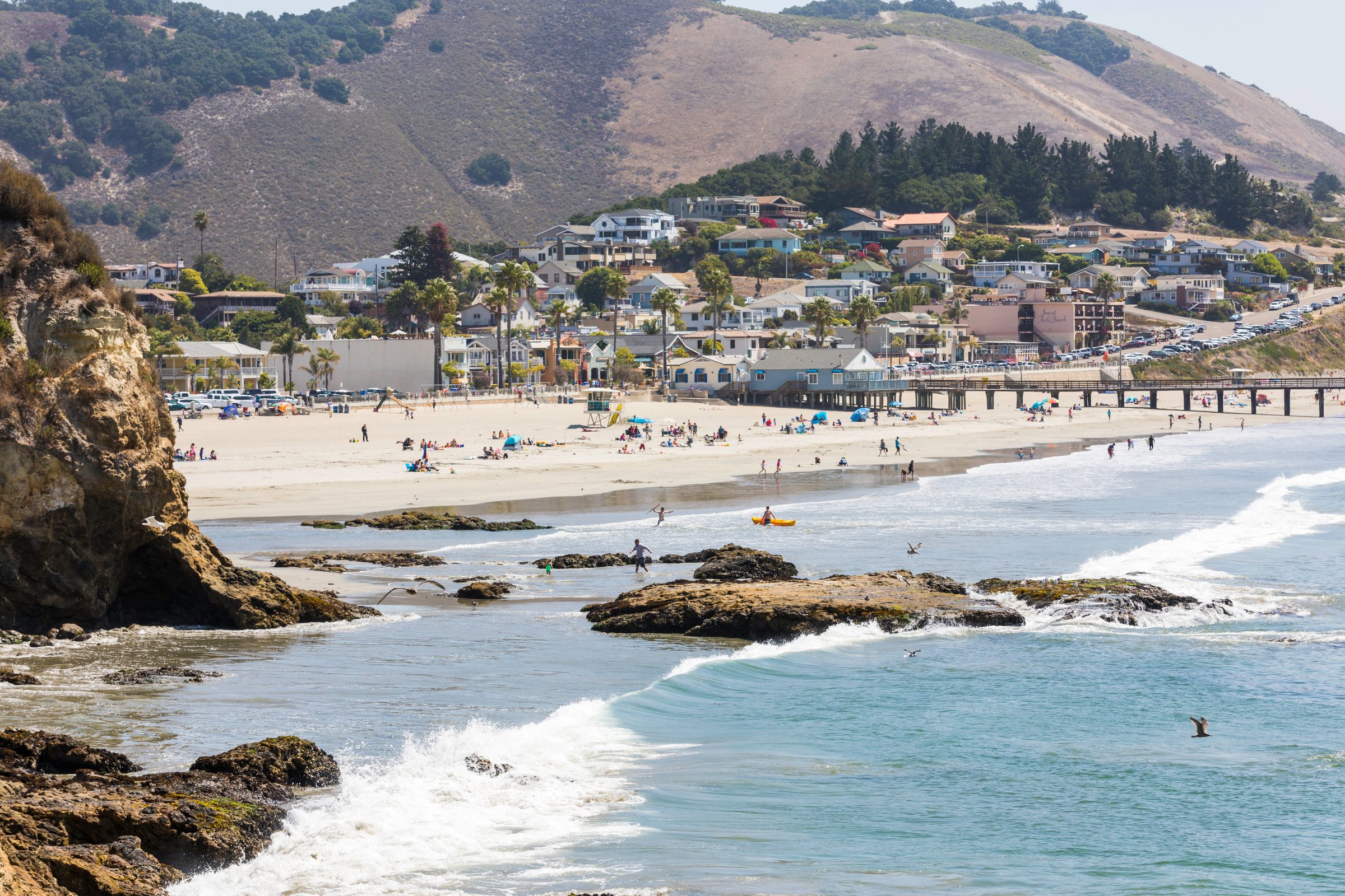 Waves washing over the rocks at the east end of Avila Beach, with the busy summer beach, the town and the hills behind