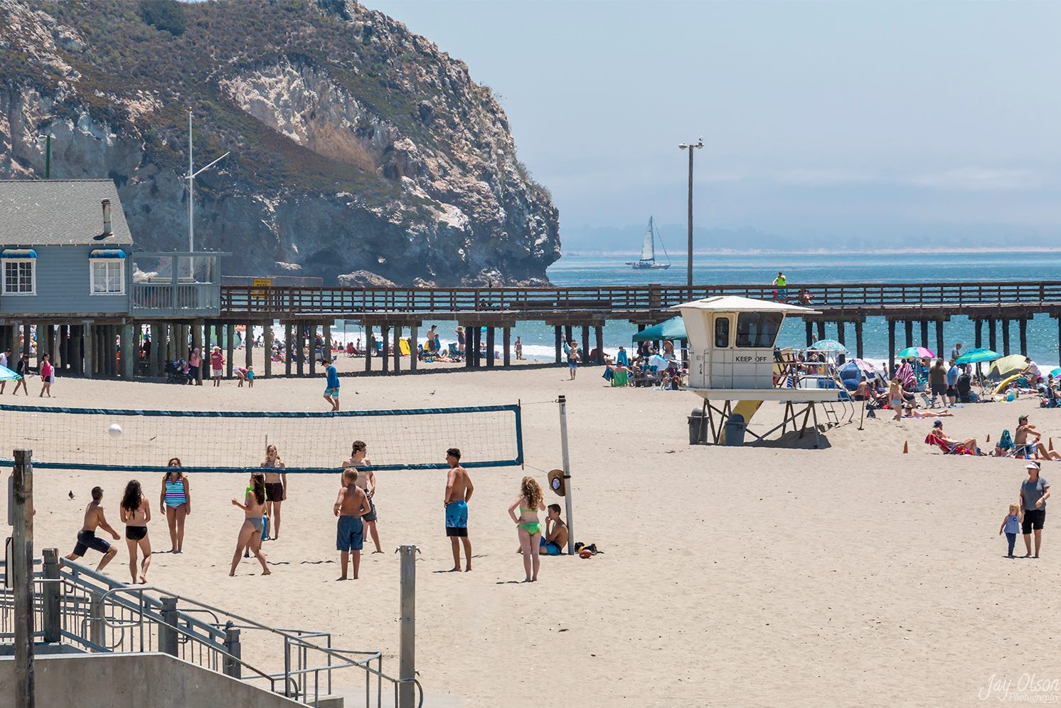 A volleyball game on a busy summer day at Avila Beach, with the pier and headland behind