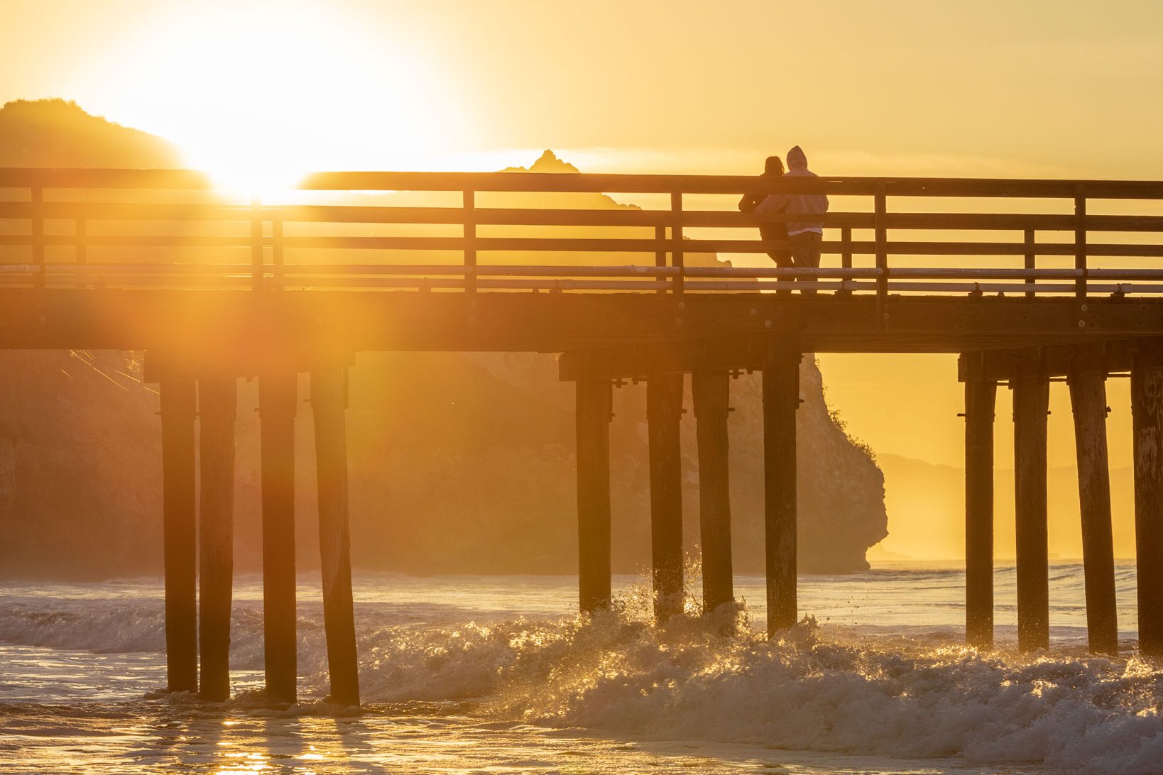 A couple watching the sunrise from Avila Pier as waves wash through the pilings