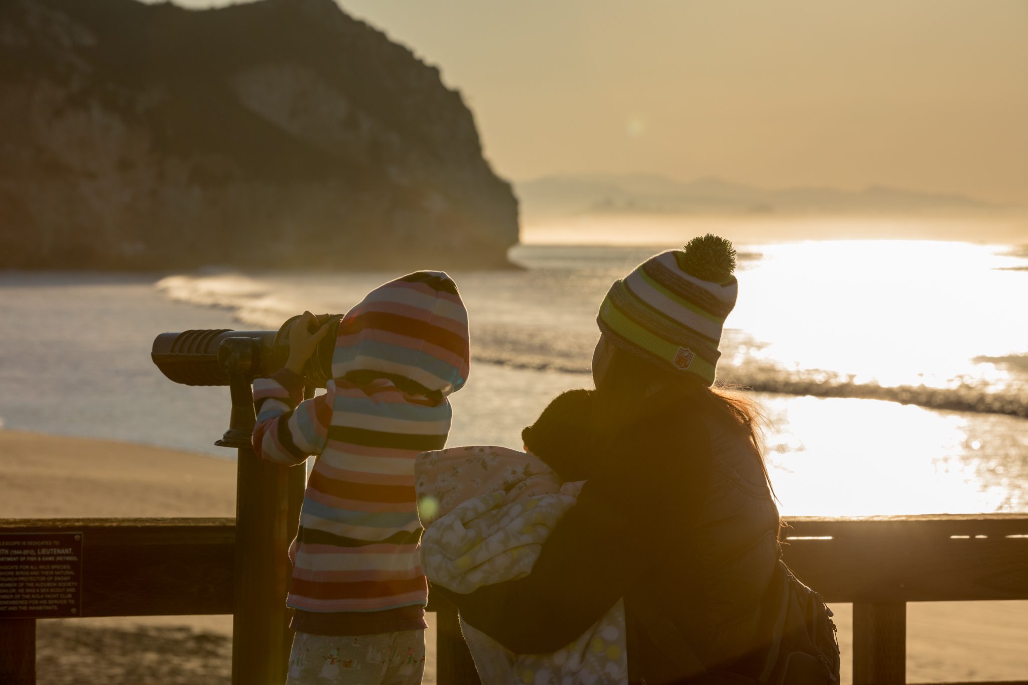 A child in a striped hoodie looking through the pier telescope at sunrise beside a parent holding a baby
