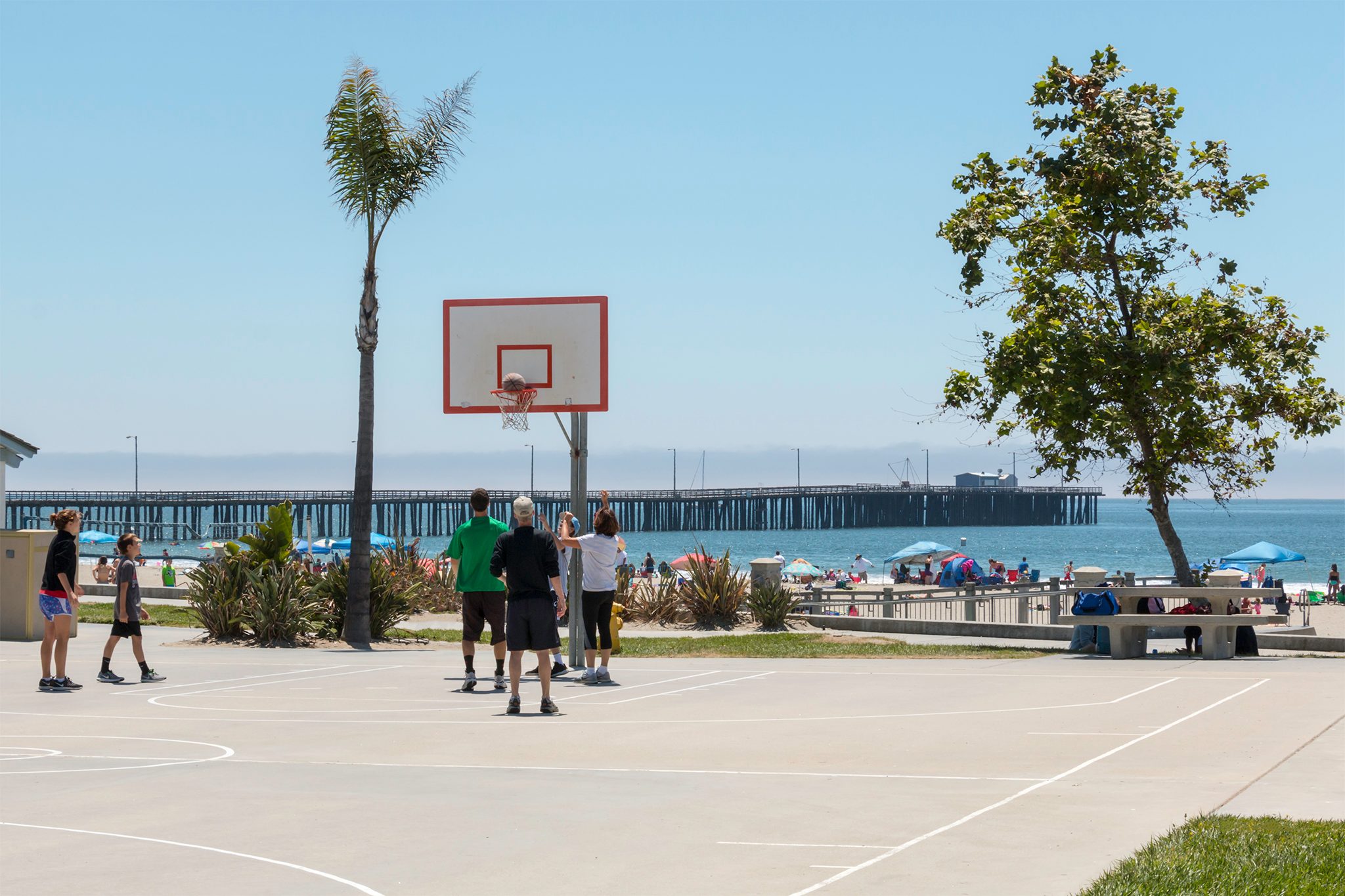 A pickup basketball game at Avila Beach Park, with the beach and pier behind
