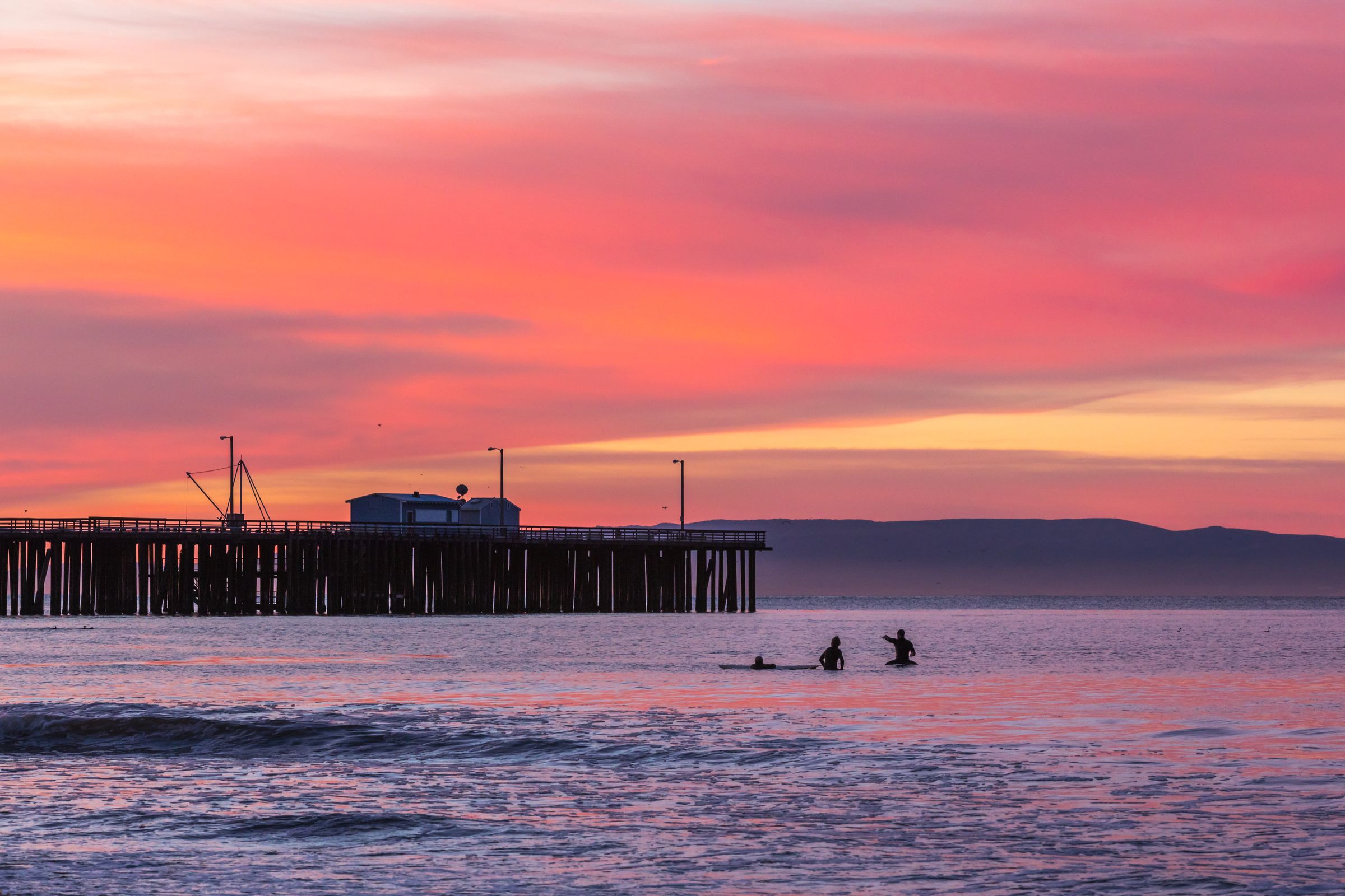 Surfers in the water beside Avila Pier under a pink sky