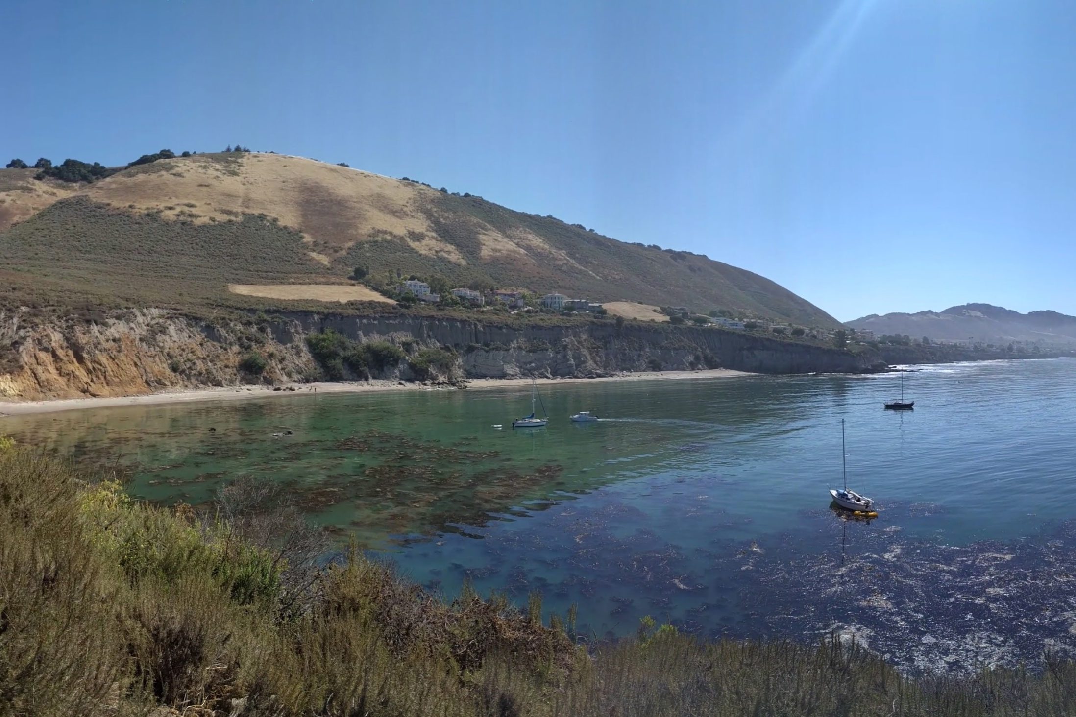 Pirate's Cove from the bluff trail, with sailboats anchored in clear green water below the hills and the beach