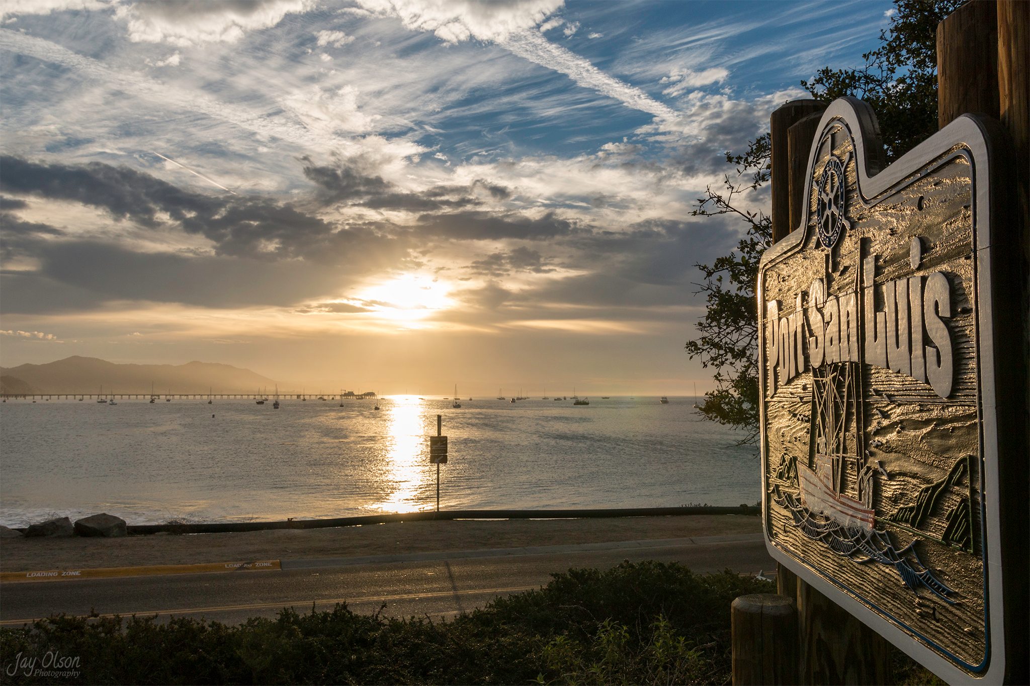 The carved Port San Luis sign above the bay, with Avila Pier and moored boats in the morning light