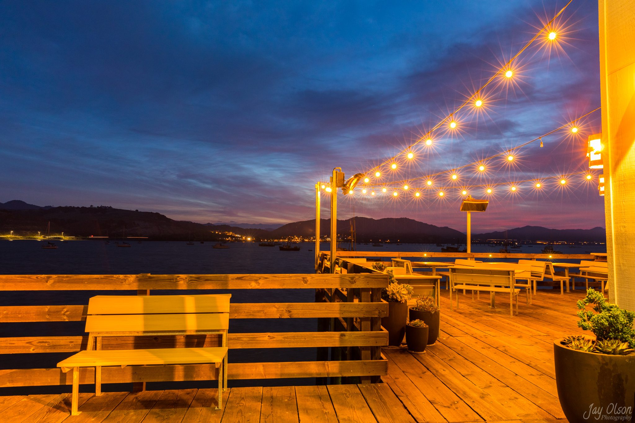 An empty restaurant deck under string lights on Harford Pier at dusk, boats moored in the bay