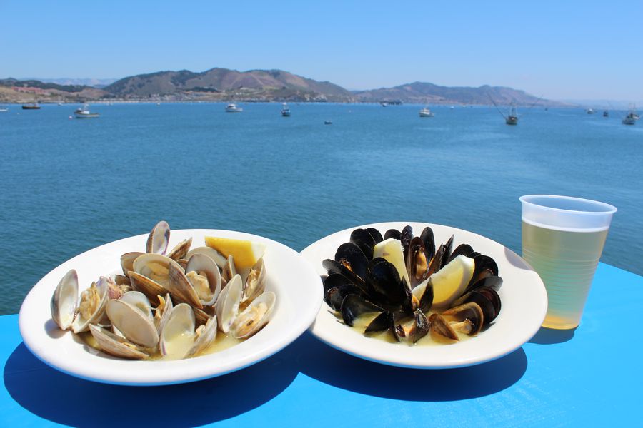 Bowls of steamed clams and mussels with lemon and a beer on a blue table over San Luis Bay, with boats and the hills behind