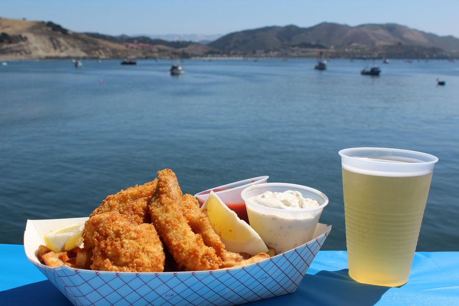 Fish and chips with tartar sauce and a cold beer on a railing at Port San Luis, with boats moored in the bay behind