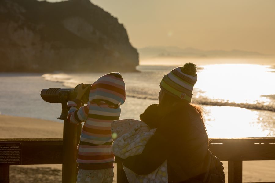 A child in a striped hoodie looking through the pier telescope at sunrise beside a parent holding a baby