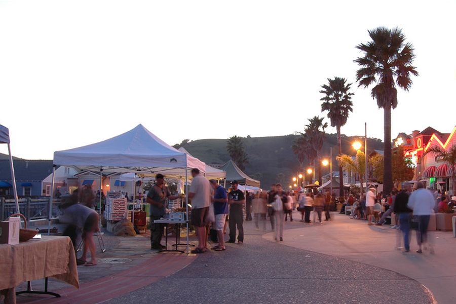 Market stalls and shoppers along the promenade in the evening light, with palms and the hills behind
