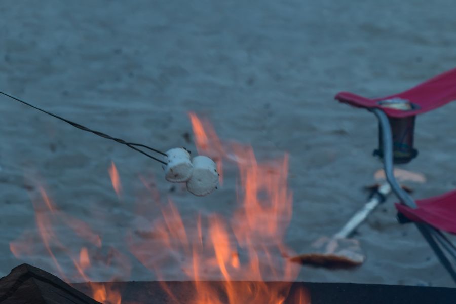 Toasting marshmallows over a fire ring on the sand at Olde Port Beach, with a beach chair and the bay behind