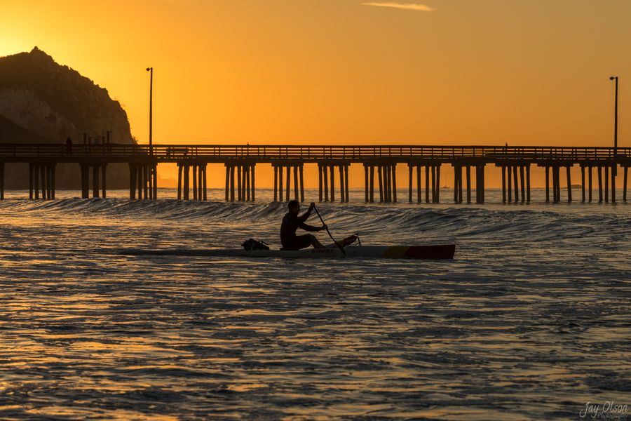A paddler in an outrigger canoe in front of Avila Pier under an orange sunrise sky
