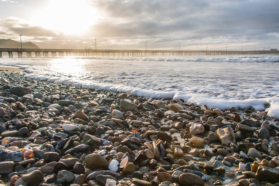 Wet pebbles and shells at the water's edge with Avila Pier under a cloudy sky