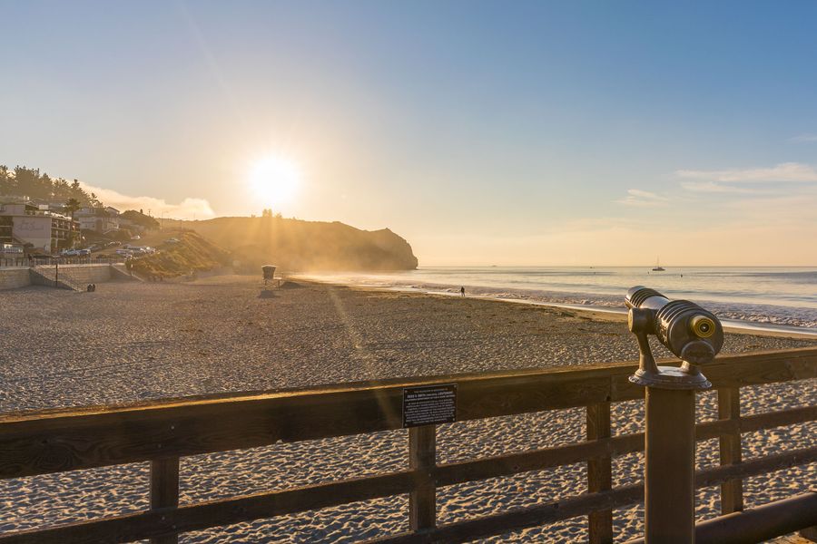 A telescope on the Avila Pier railing looking over the beach toward the headland at sunrise