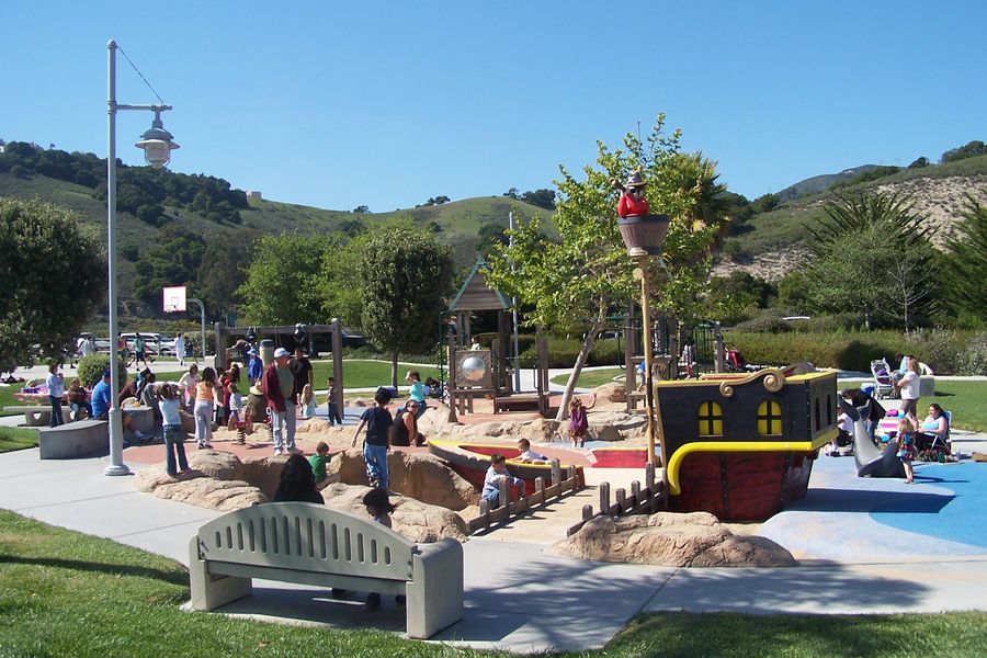 Families at the pirate ship playground in Avila Beach Park, with a sand pit, climbing rocks, a basketball hoop and green hills behind