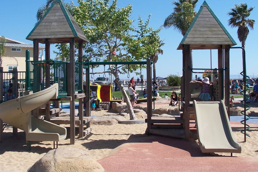 Slides and climbing towers in the sandy playground at Avila Beach Park, with palms and the promenade behind