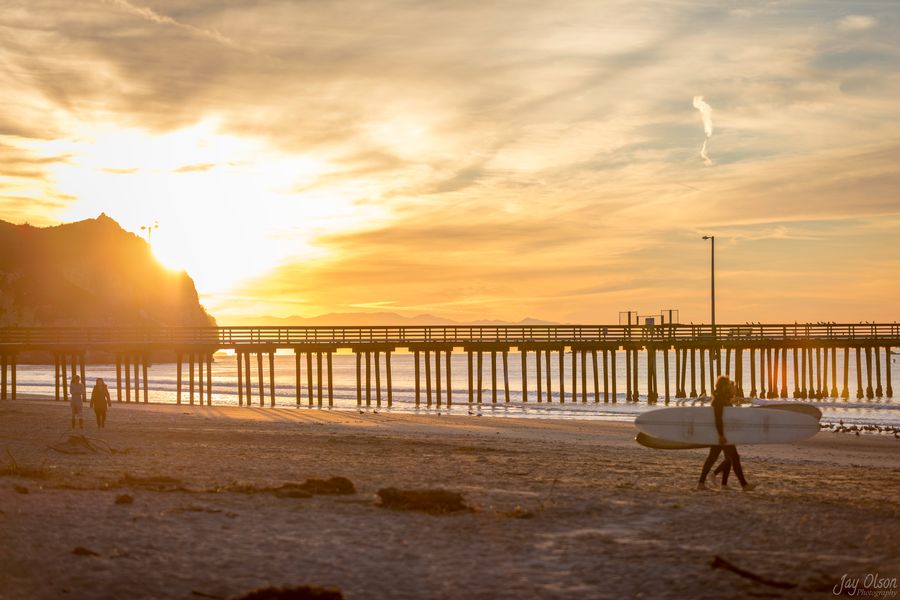 Two surfers carrying longboards past Avila Pier under a golden sunrise sky