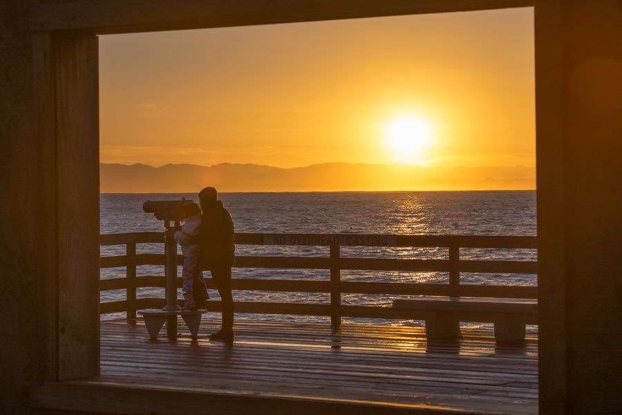 A parent and child at the pier telescope, framed by a window at sunrise