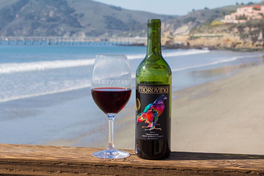 A glass of red wine and a bottle on a railing above Avila Beach, with Harford Pier behind
